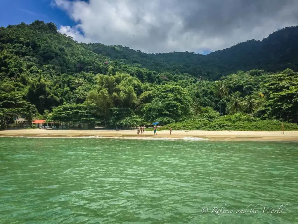 Secluded beach on Ilha Grande, Brazil, with a few visitors on the sandy shore, surrounded by lush, dense tropical forest and clear green waters. Enjoy the gorgeous beaches in Ilha Grande, Brazil.