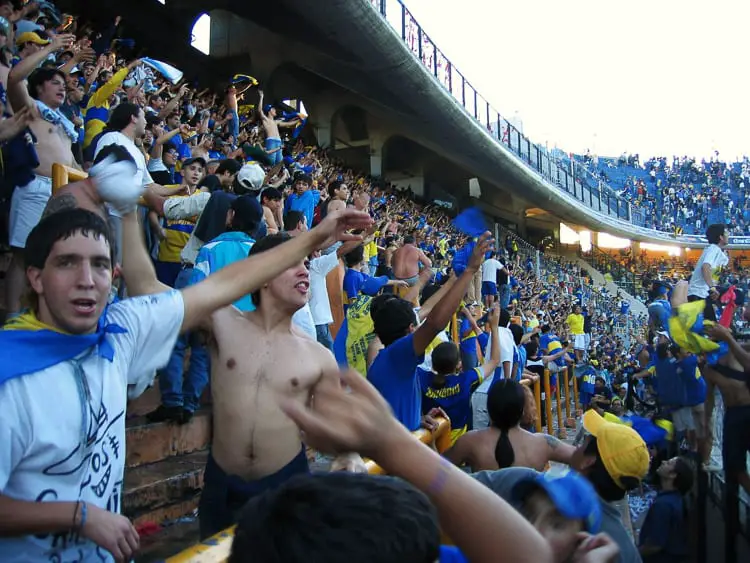A dynamic scene of enthusiastic Boca Juniors football fans filling the stadium stands, waving flags, cheering, and clapping in a sea of yellow and blue. The energy of the crowd is palpable, showcasing the passionate spirit of Argentine football culture. Visiting a football match in Buenos Aires is a crazy experience!