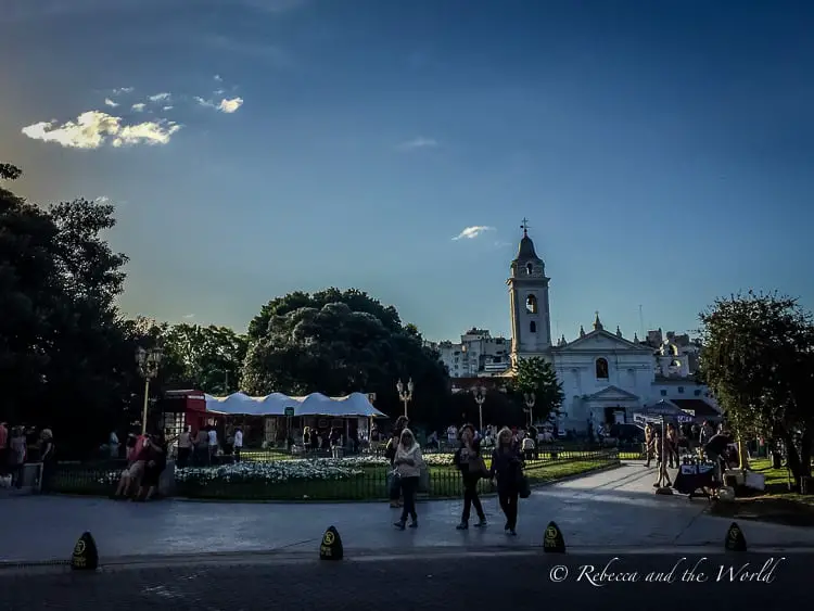 Visitors milling around a bustling outdoor market in a park in Buenos Aires, with the historic white church and clock tower of Nuestra Señora del Pilar in the background. The Basilica de Nuestra Senora del Pilar is a gorgeous white church in Recoleta, a neighbourhood of Buenos Aires.