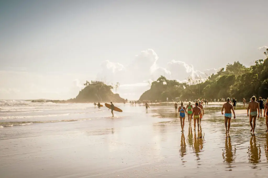 Beachgoers walking along a shoreline, with surfers carrying their boards and a rock island in the distance under a cloudy sky. A road trip up the east coast of Australia is one of the best road trips in Australia.