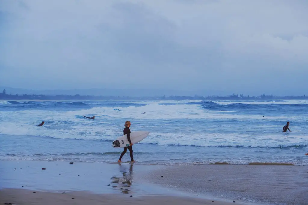 A surfer walking along a stormy beach with waves rolling in, under a moody, overcast sky. Byron Bay is one of the best places to visit in Australia for its laid-back surf vibe.