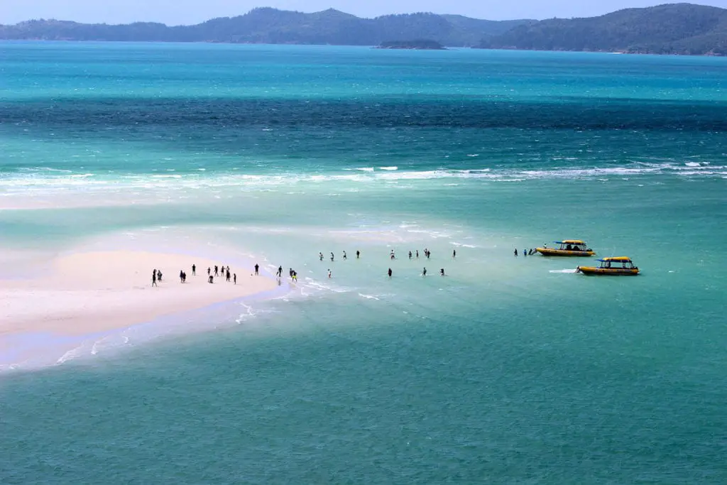 Beachgoers walking on a sandbar with clear turquoise water, boats nearby, and a forested island in the distance. The Whitsunday Islands are some of the most beautiful islands in Australia. Just off the coast of Queensland, you can visit them on a sailing trip.