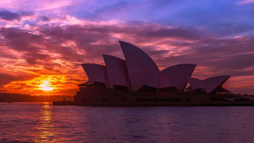 Sydney Opera House silhouetted against a vibrant sunrise, with rich orange and purple hues reflecting on the water. The Sydney Opera House is one of Australia's most recognisable buildings and a tour of its interior is one of the best things to do in Sydney.