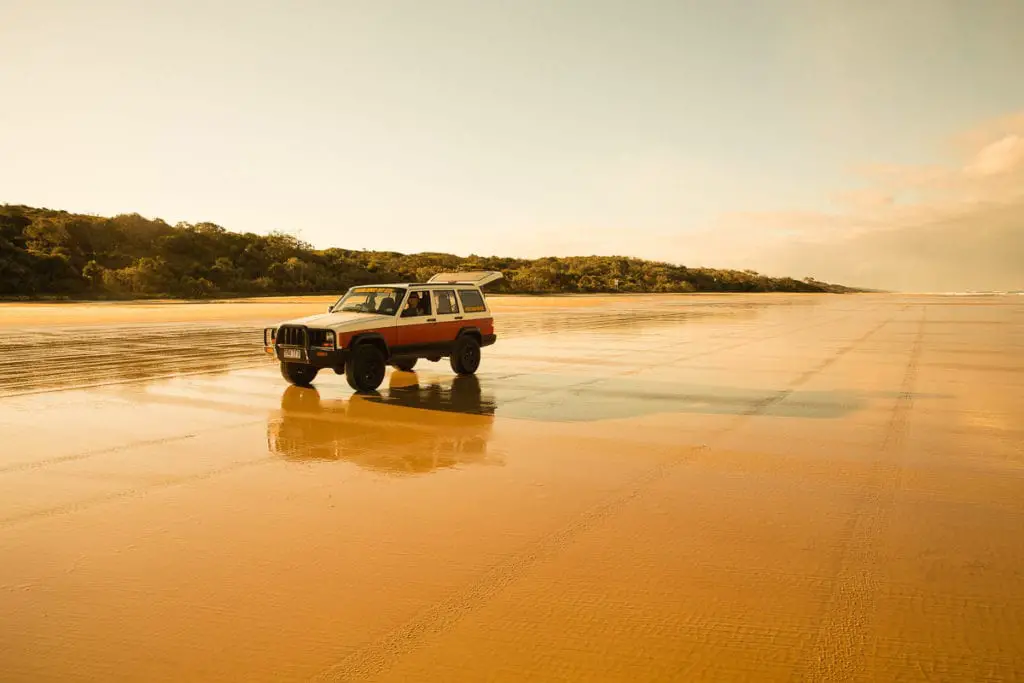 A four-wheel drive vehicle on a wide sandy beach with wet sand reflecting the sunlight, surrounded by a distant forest. K'gari (formerly called Fraser Island) is the world's largest sand island and one thing for your Australia bucket list is to go four-wheel driving on the sand!