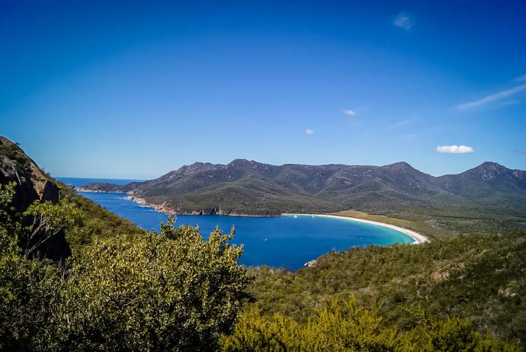 Panoramic view of Wineglass Bay with turquoise waters surrounded by green hills under a blue sky. Wineglass Bay is just one of Tasmania's beautiful coastlines.