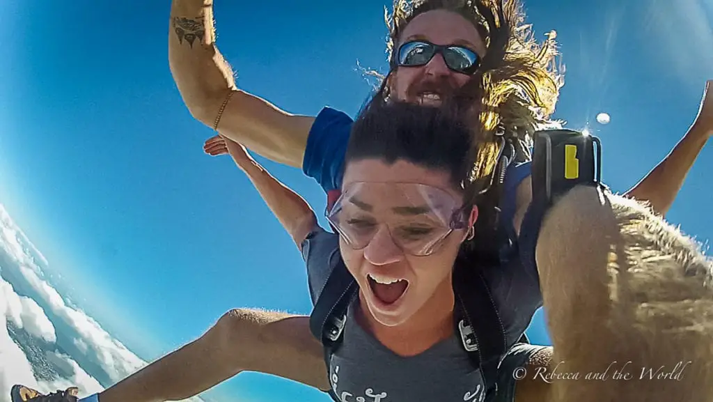 Two people tandem skydiving, the skydiver in the foreground - the author of this article - is screaming with excitement while the instructor behind is holding out arms, against a backdrop of clear blue sky and clouds. You can skydive at many places in Cairns, Australia (although don't expect to look good in photos doing it!).