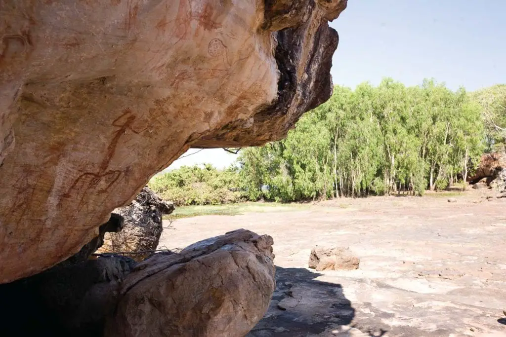 Ancient rock art on the underside of a rock shelter in a dry Australian landscape with greenery in the background. Arnhem Land is an incredible place to visit in Australia to learn about Aboriginal Australia history.