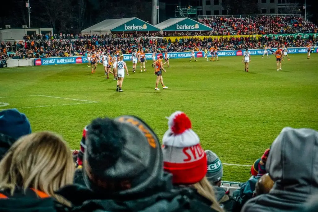 Spectators at an Australian rules football game, watching intently from the stands with players in the background. AFL is a sport in Australia that's a must-watch when you visit the country.