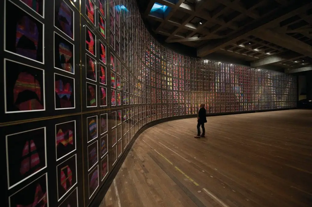 A large, darkened gallery wall displaying an array of colorful square images, with a lone person standing in the corner for scale. The Museum of Old and New Art is one of the most talked-about museums in Australia.