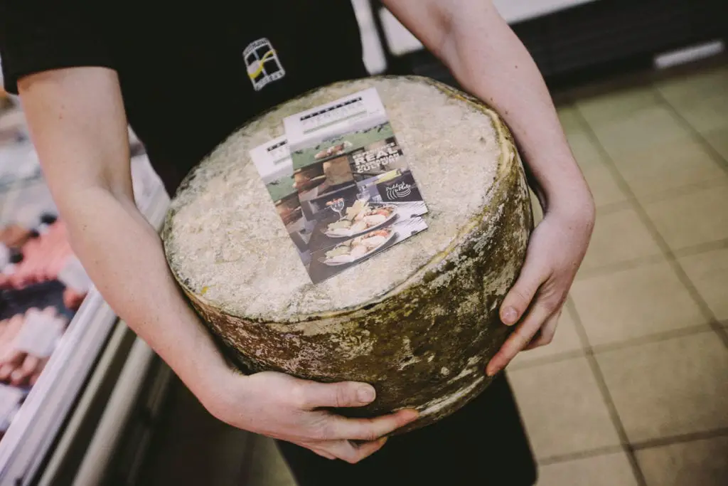 A person holding a hefty wheel of cheese with an informational card on top, indicating a local Australian produce. Tasmania is known for its incredible cheeses - tasting local products is just one of the many things to do in Tasmania, Australia.