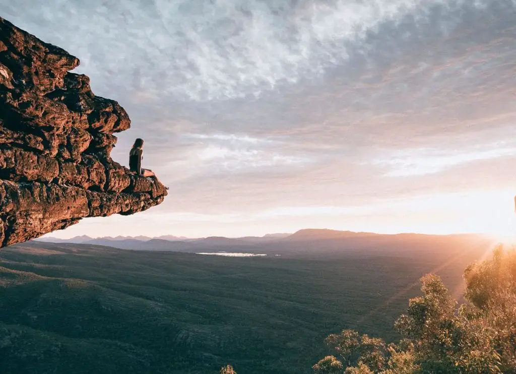 A solitary person sitting on the edge of a high cliff overlooking a vast wilderness at sunset. The Grampians is a beautiful place to visit in Australia, with plenty of hiking, wildlife spotting and panoramic views.