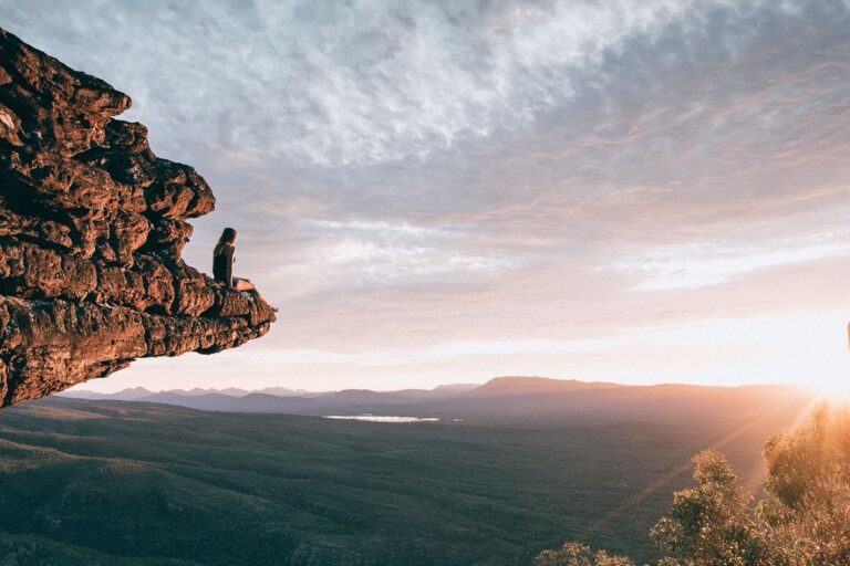 A solitary person sitting on the edge of a high cliff overlooking a vast wilderness at sunset. The Grampians is a beautiful place to visit in Australia, with plenty of hiking, wildlife spotting and panoramic views.