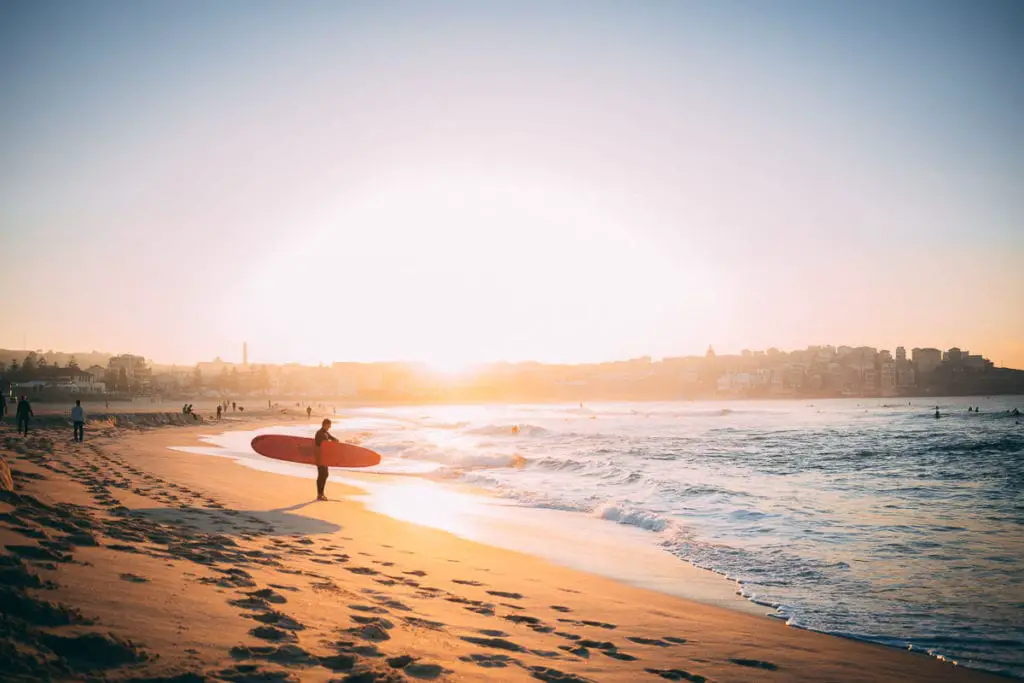 Silhouette of a surfer holding a red board on a beach at sunset, with the ocean's horizon line and surfers in the water. Bondi Beach is one of the most famous beaches in Australia.