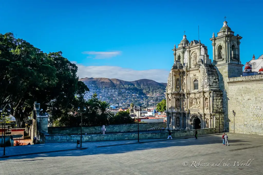 A spacious plaza with a large, ornate church featuring two bell towers and an intricate facade. The plaza is bordered by trees, and a few individuals are scattered across the wide, open space. Hills are visible in the distance under a bright blue sky. The views and architecture in Oaxaca are stunning.