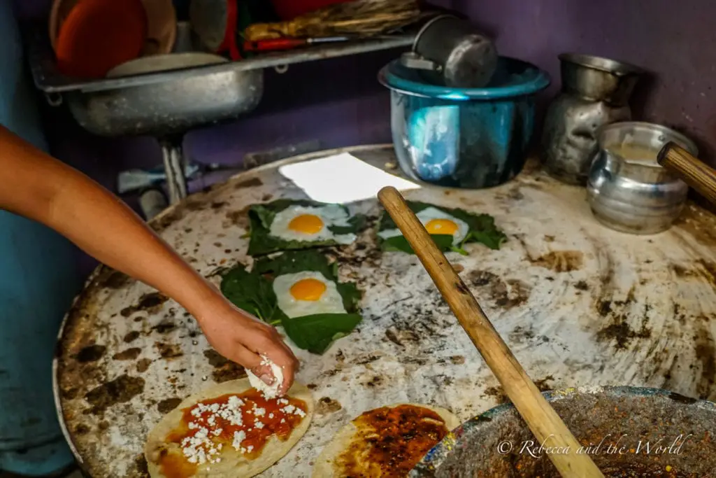 An individual preparing food on a large, round, stone surface. Fresh tortillas are being topped with sauce and cheese, and there are leaves with eggs on them. Cooking utensils and pots are arranged around the preparation area. The food in Oaxaca is incredible - plan your Oaxaca itinerary around trying all the specialties of the region.