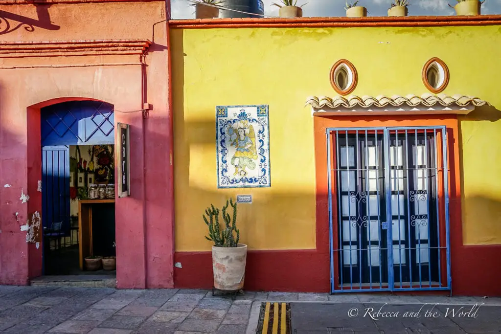 A vibrant section of a building painted yellow with red trim. A blue door is open, leading into a dimly lit interior, and a blue-framed window with iron bars is closed. A decorative tile with an image of a lion is on the wall next to a potted cactus. Oaxaca has several colourful buildings and homes.