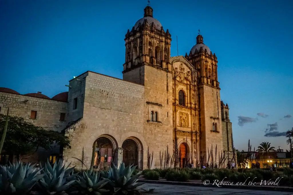 A majestic church captured at dusk, its ornate facade and twin bell towers illuminated against the twilight sky. The church stands next to a serene courtyard with agave plants in the foreground. The Templo de Santo Domingo is a Baroque church that's stunning to visit - it's one of the best things to do in Oaxaca.