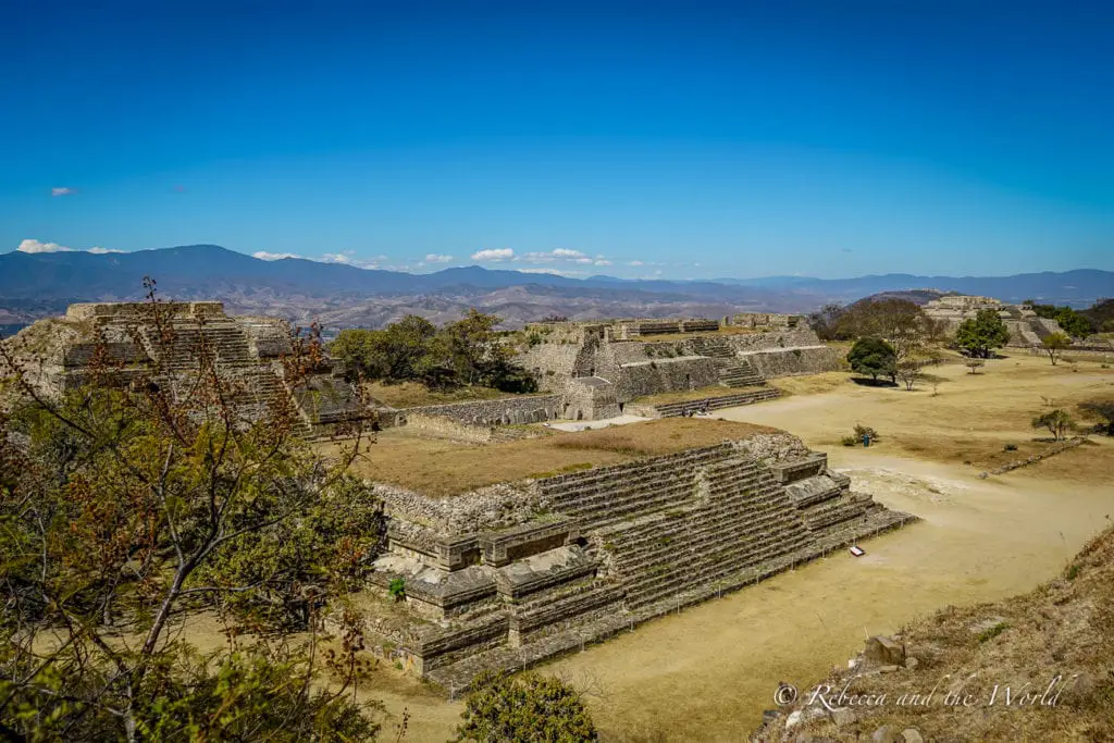 An ancient Mesoamerican archaeological site with several step pyramids and structures. The ruins are surrounded by a sparse landscape with distant mountains under a clear sky. Visiting Monte Alban is one of the best things to do in Oaxaca to learn about pre-Columbian history.