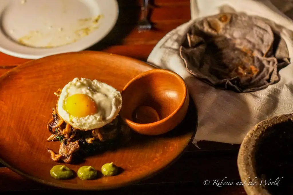 A gourmet dish consisting of a base of sautéed mushrooms topped with a fried egg, accompanied by a few dollops of green sauce. The plate is wooden, and there's a cloth napkin with a tortilla on the table. Oaxaca food is delicious and Casa Oaxaca showcases local ingredients in their dishes.