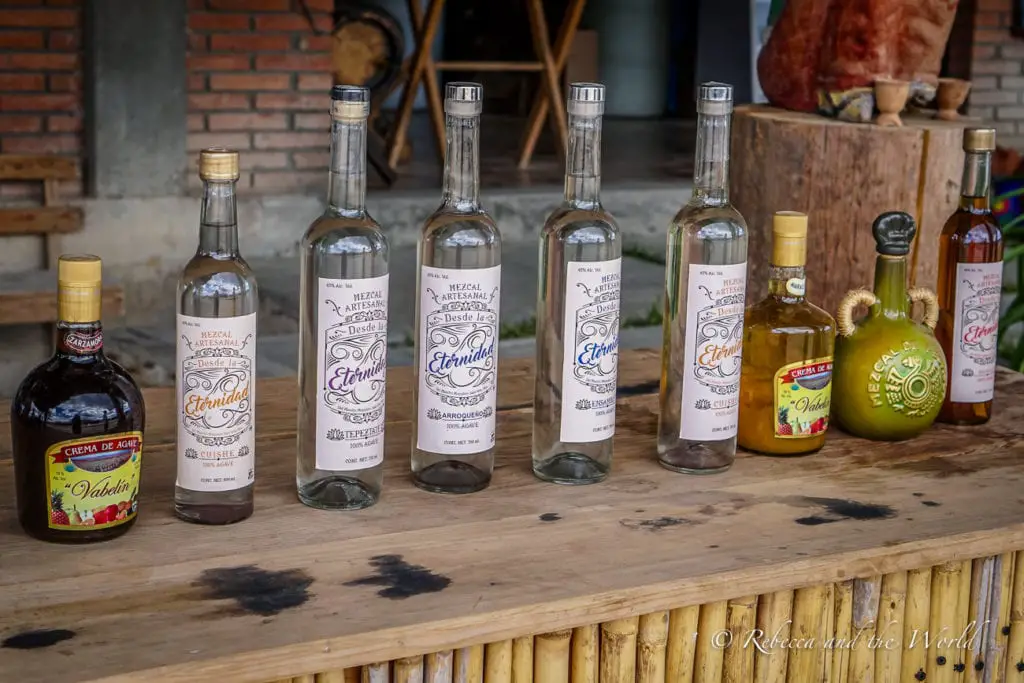 A series of clear glass bottles labeled "Mezcal Artesanal" are lined up on a wooden counter. Each bottle has a different label design, and they are accompanied by two dark bottles of "Crema de Agave". A mezcal tour is one of the best things to do in Oaxaca to discover how this liquor is made.