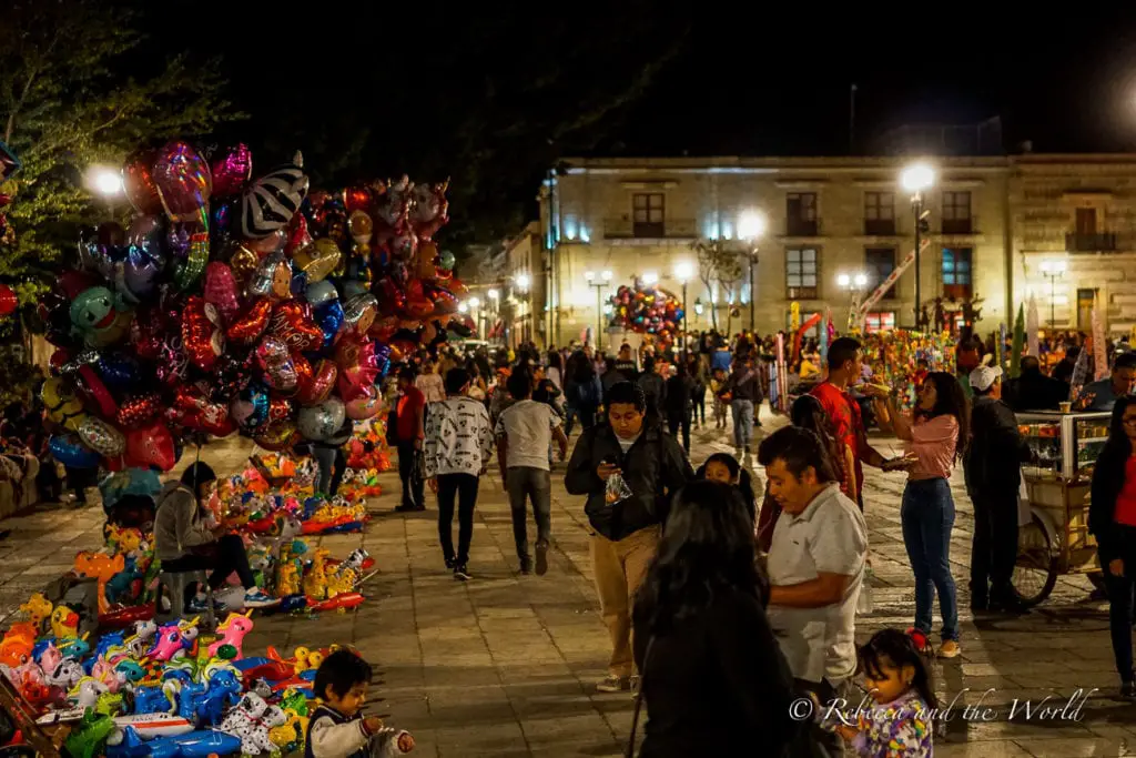 A bustling night market scene with a crowd of people walking between vendors. Colorful balloons and toys are for sale, creating a vibrant atmosphere. The market is lit by streetlights and takes place in an open plaza setting. The Zocalo in Oaxaca is a fun place to hang out at night time.