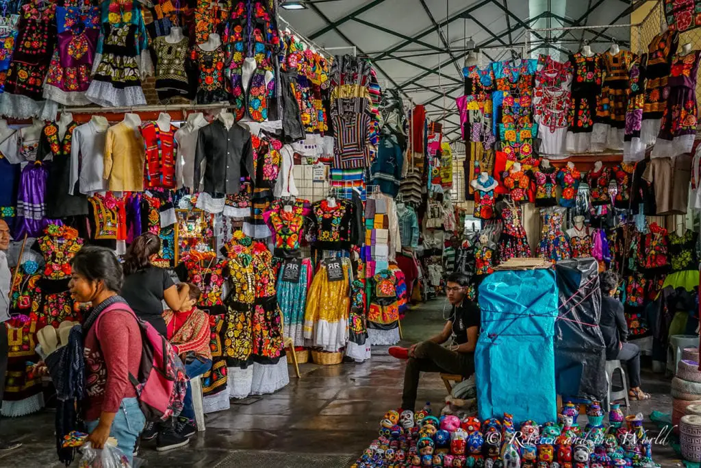 An indoor market scene with stalls draped in a variety of colorful textiles and garments. People browse through the merchandise, and the atmosphere is busy and vibrant with traditional clothing and crafts on display. Pack a spare bag when you visit Oaxaca - the shopping is phenomenal.