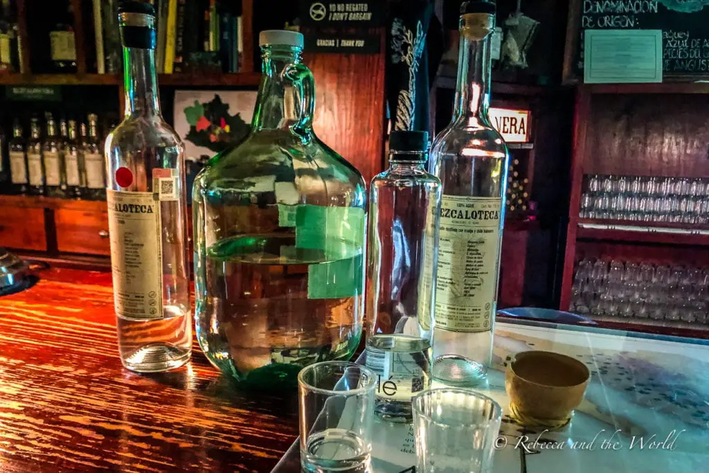 A dimly lit bar scene showcasing bottles of mezcal on a wooden counter. The bottles have labels providing details about the spirit, and there are empty glasses and a clay cup nearby, suggesting a tasting in progress. Try mezcal, a specialty of the Oaxaca region, at La Mezcaloteca.