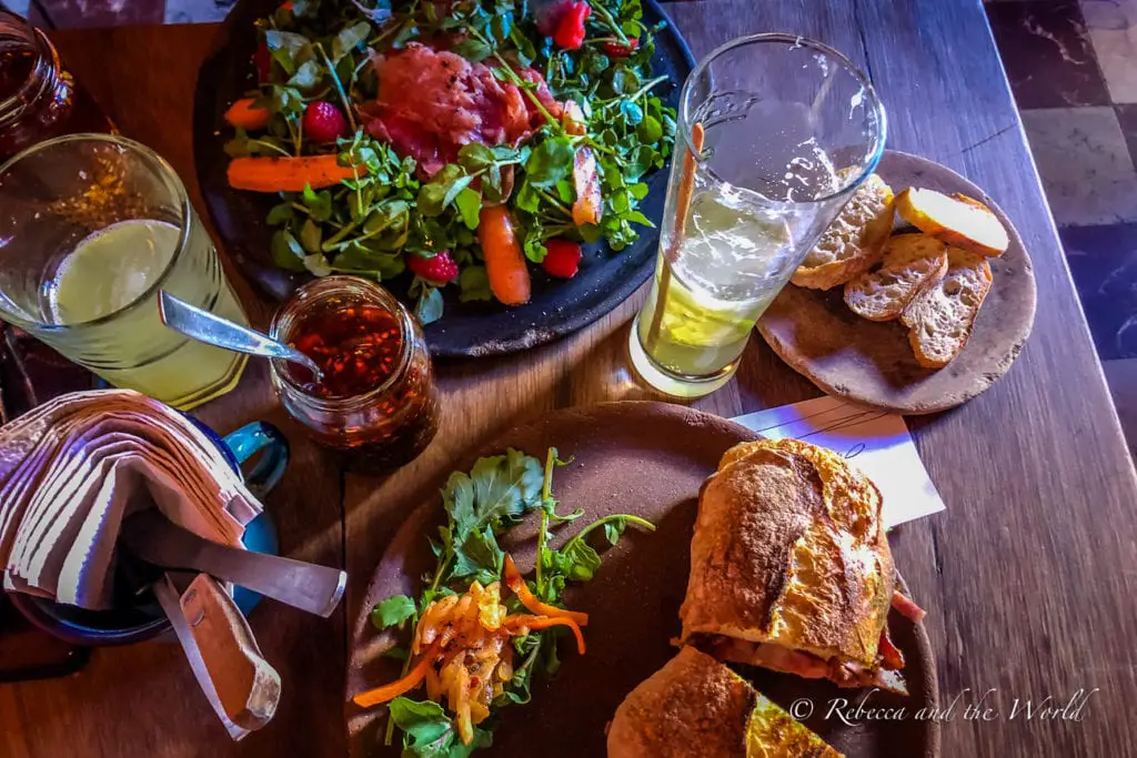 A meal laid out on a wooden table, consisting of a vibrant salad, a sandwich cut in half with visible fillings, and two glasses of a yellowish drink. A jar of red chili flakes and a folded wallet are also on the table. Lunch at Boulenc should be on your Oaxaca itinerary.