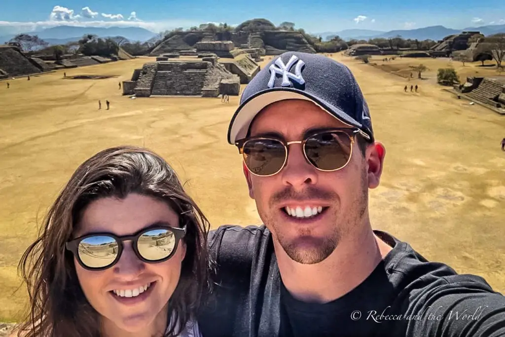 A selfie of two smiling people - the author and her husband - wearing sunglasses. In the background, the ruins of an ancient Mesoamerican city stretch out, with several pyramids visible under a blue sky. Visiting Monte Alban is one of the best things to do in Oaxaca to learn about pre-Columbian history.