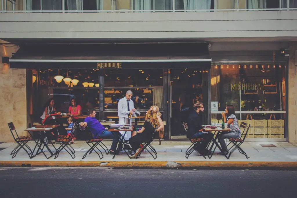 An outdoor restaurant scene with patrons seated at tables served by a waiter, with the restaurant's name visible above the entrance - Mishiguene. Mishiguene is one of the best places to eat in Buenos Aires, Argentina.