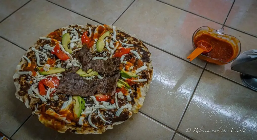A traditional Oaxacan tlayuda topped with beans, cheese, avocado, tomato, and steak, accompanied by a side of salsa. Oaxacan food is incredible, including tlayudas, which is often called a Mexican pizza.