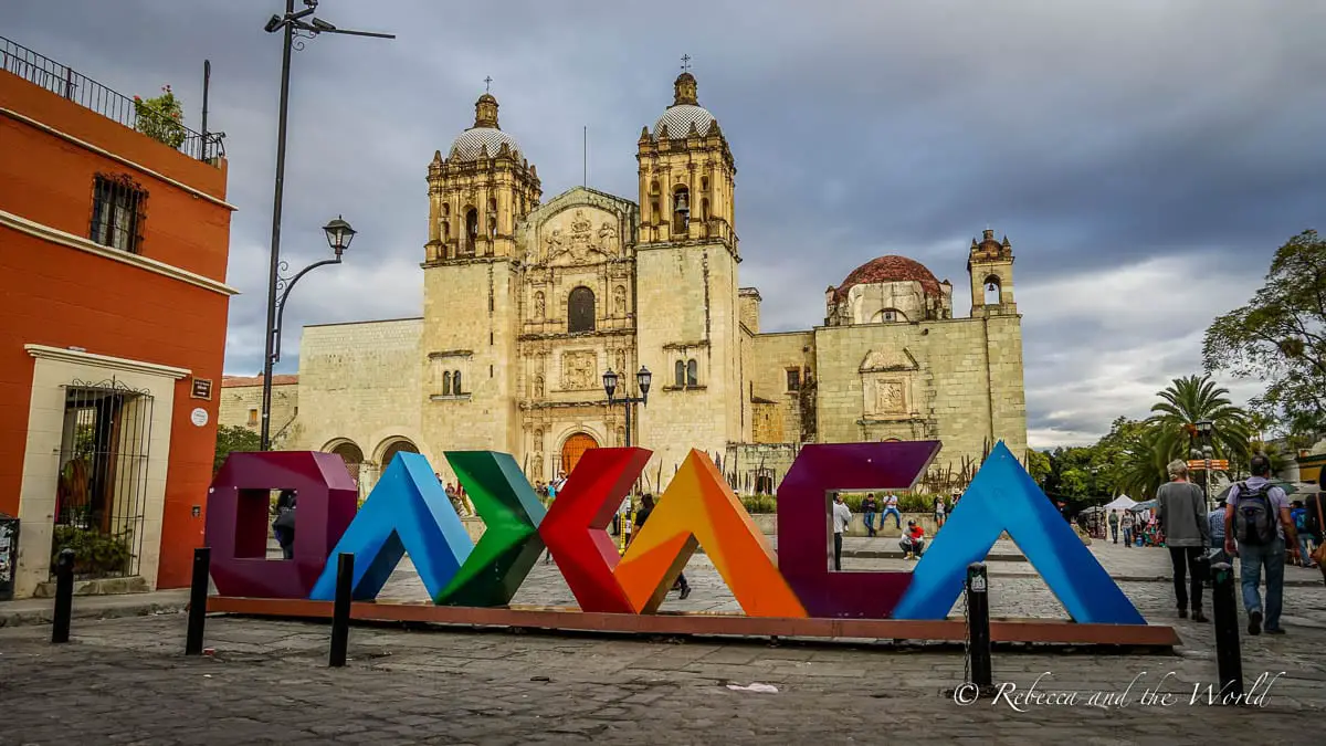 Colorful 'OAXACA' sign in bold letters with the Santo Domingo de Guzmán church in the background under a cloudy sky. Oaxaca is one of the best places to visit in Mexico, but it can be tricky to pronounce!