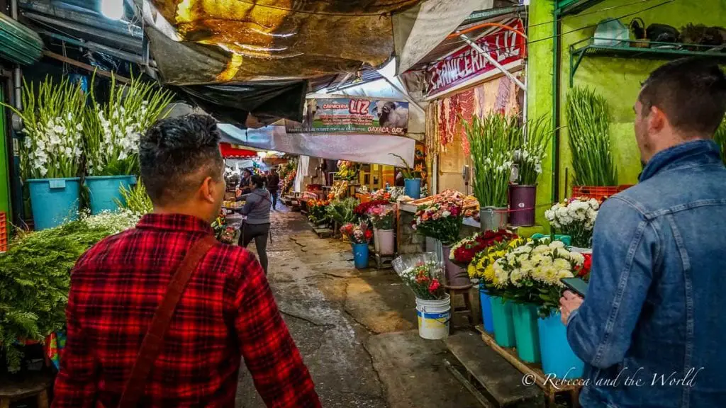 Busy market scene in Oaxaca with stalls lined with fresh flowers and people walking through. If you're travelling to Oaxaca, consider a food tour with Omar of Oaxacking to learn what, where and how to eat Oaxacan food.
