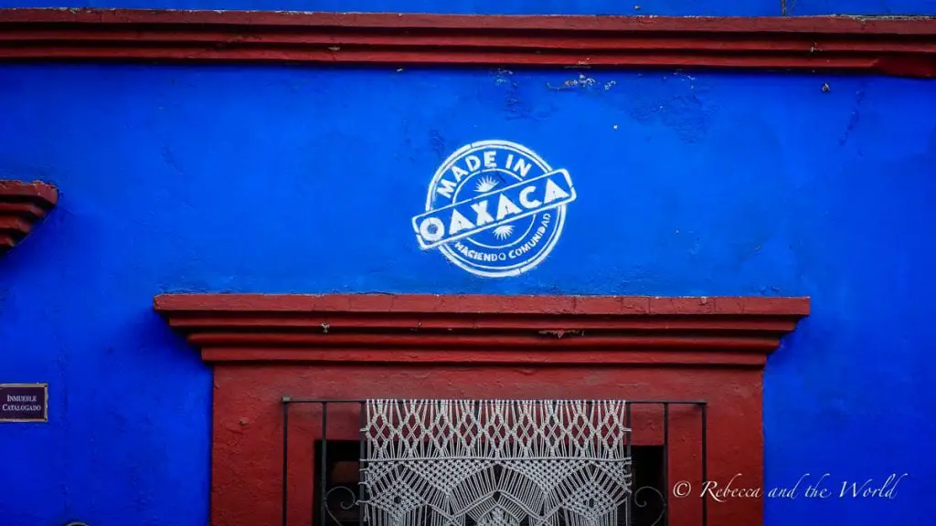 Vibrant blue wall with 'MADE IN OAXACA - HACIENDA COMUNITARIA' emblem painted in white, with red trim and a decorative white iron window grille. Oaxaca travel is super easy to organise - there's an international airport in the city, or you can fly via Mexico City.
