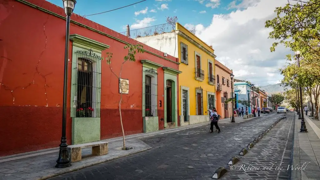A street in Oaxaca lined with colorful buildings and cobblestone pavement, pedestrians walking on the sidewalk. One of the things to know before you visit Oaxaca is that the city is meticulously clean and well-kept.