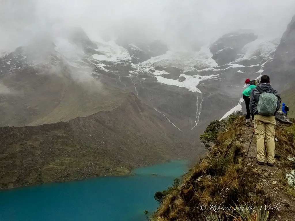 Hikers observing a deep blue glacial lake from a narrow mountain trail, with mist covering the higher peaks in the Salkantay region.