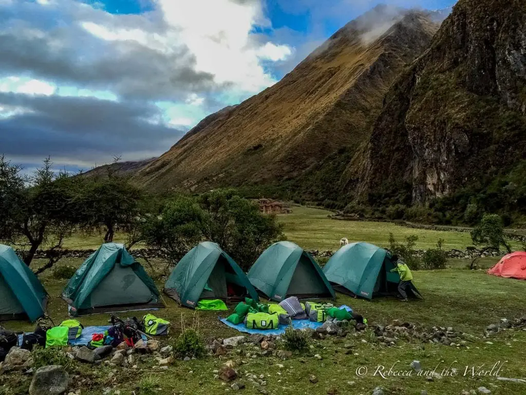 Campsite with several green tents set up on a grassy field with mountains in the background under a cloudy sky on the Salkantay Trek. Hiking the Salkantay Trek to Machu Picchu in Peru means some photogenic camping spots along the way.