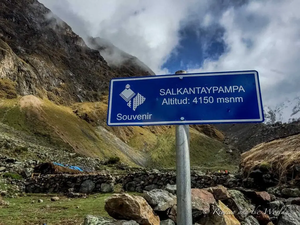 Blue directional sign reading 'SALKANTAYPAMPA Altitud: 4150 msnm' with mountains in the background and a small stone hut nearby. The Salkantay Trek is high-altitude hiking, so make sure to pack the right medications and come prepared.
