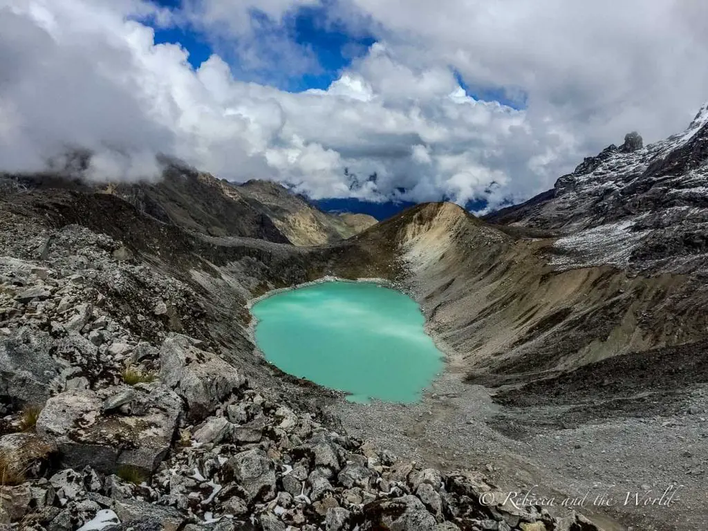Turquoise glacial lake surrounded by rocky mountains with patches of snow, under a partly cloudy sky. There are several blue and green lakes along the Salkantay Trek to Machu Picchu.