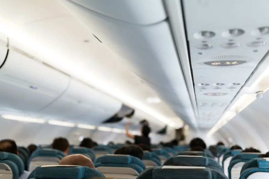 The interior of an airplane looking towards the front of the cabin. The overhead compartments are closed, and the cabin has rows of blue seats occupied by passengers. The focus of the image is on the airplane ceiling, with overhead lighting and air conditioning nozzles. A flight attendant is standing in the aisle, appearing to be interacting with a passenger or conducting a task.