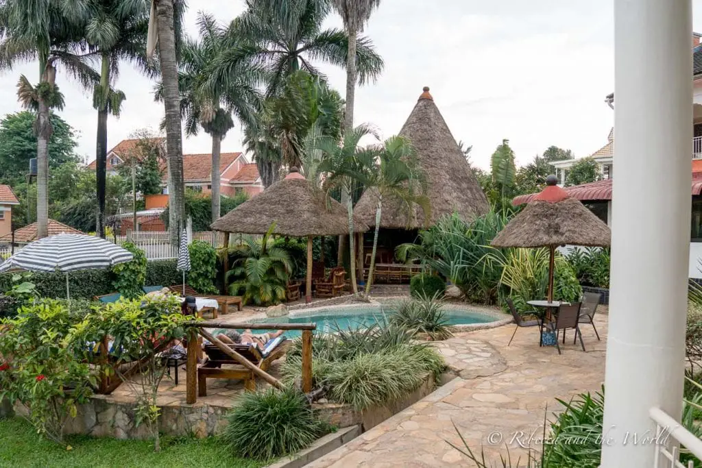 A tranquil poolside setting with thatched umbrellas, surrounded by tropical plants and palm trees. A person lounges by the pool, and residential buildings are visible in the background. 2 Friends Beach Hotel in Entebbe is a great choice if you're wondering where to stay in Entebbe, Uganda.