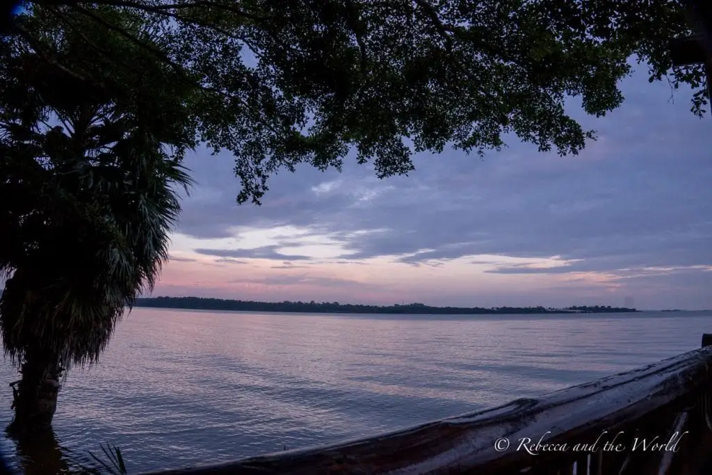A dusk view over a calm Lake Victoria in Uganda, with clouds reflecting the soft blue and purple hues of twilight. Trees and palms are silhouetted against the evening sky. 2 Friends Beach Hotel in Entebbe, Uganda, has a restaurant right on the shores of Lake Victoria.