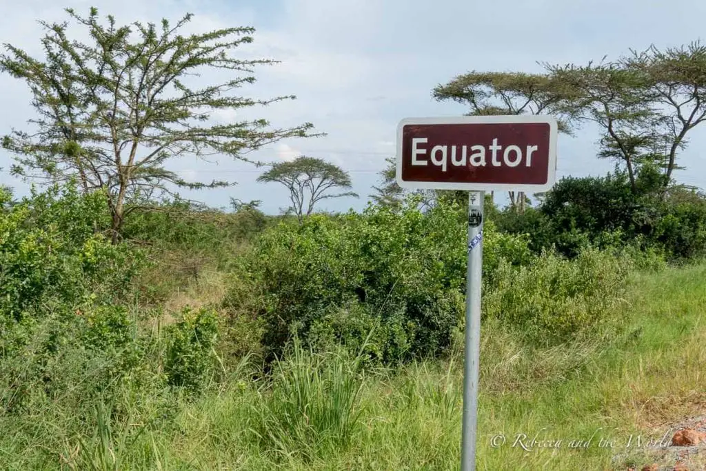 A roadside sign reading "Equator" in front of a landscape with trees and shrubs under a cloudy sky. The equator cuts through Uganda and there are signposts along major highways.