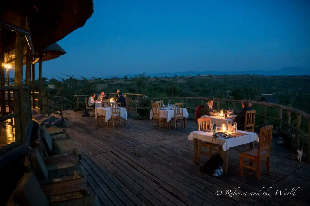 An outdoor dining area at dusk with tables set for dinner, guests seated, and lanterns providing a warm glow. The deck overlooks a vast savannah under a twilight sky. Kasenyi Safari Camp is a luxury lodge in Queen Elizabeth National Park.