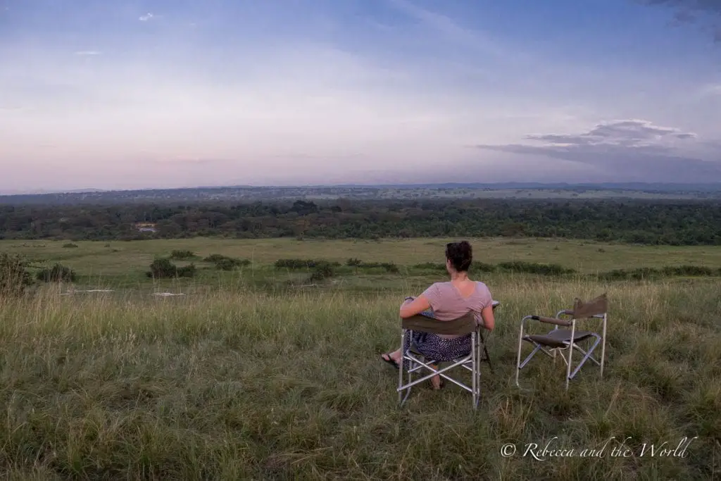 A woman (the author of this article) sits on a folding chair facing a vast African savannah under a soft, pastel-coloured sky, with an empty chair beside them, suggesting a peaceful moment in nature. A sundowner in Uganda is a must for any Uganda itinerary.