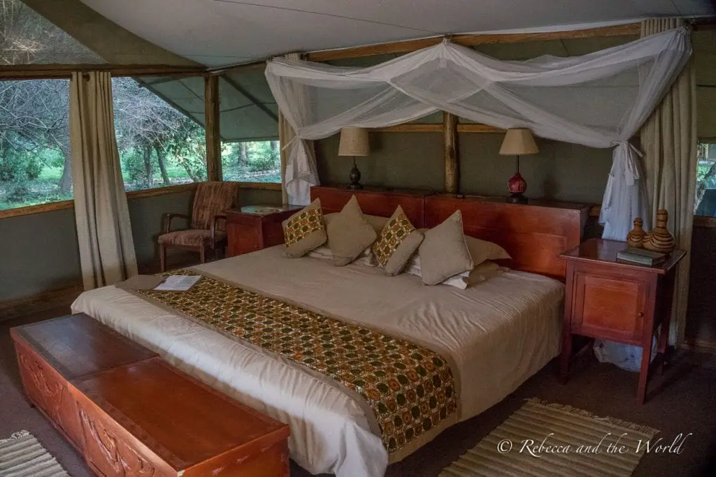 An interior view of a safari lodge bedroom with a large bed, mosquito netting, wooden furniture, and a view of trees through the canvas walls. The beds are huge at Ishasha Wilderness Camp in Uganda.