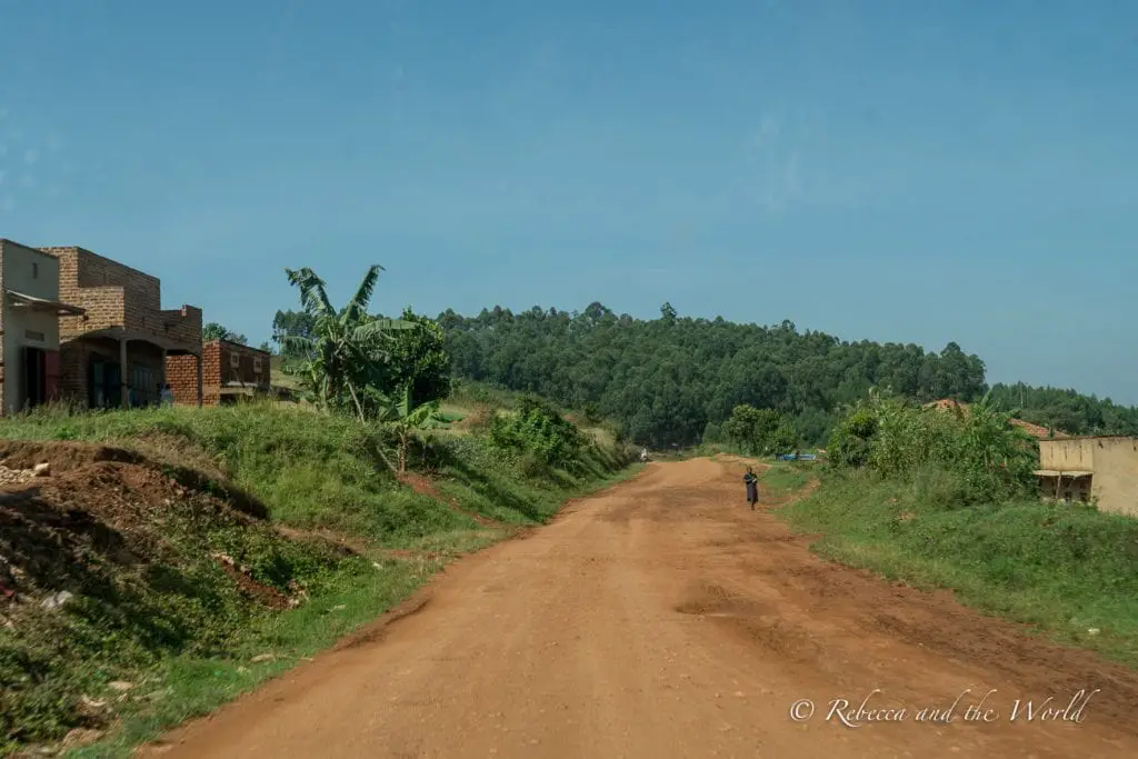 A rural scene in Uganda, seen through a car window, featuring a dirt road stretching into the distance, flanked by greenery and unfinished brick buildings. A lone figure walks down the road, and lush hills rise in the background. The best time to visit Uganda is during the dry season when road conditions will be better.