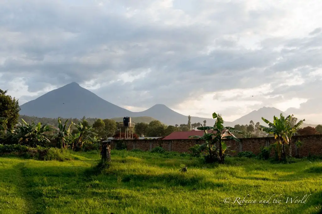 A rural landscape with banana plants in the foreground and twin volcanic mountains in the distance, under a cloudy sky. Behind the Travellers Rest Hotel in Kisoro, Uganda, you can catch a glimpse of the Virunga Mountains.