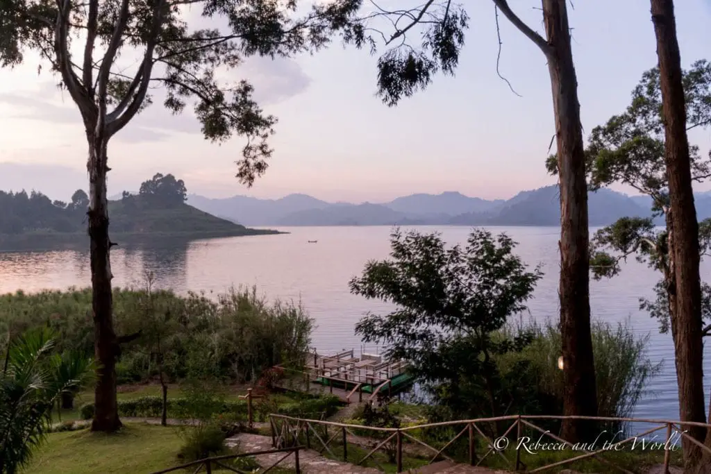 A tranquil view of Lake Mutanda at twilight, with silhouettes of trees and distant hills reflecting on the calm water surface, and a small boat visible in the distance. Mutanda Lake Resort is one of the best places to stay in Uganda.