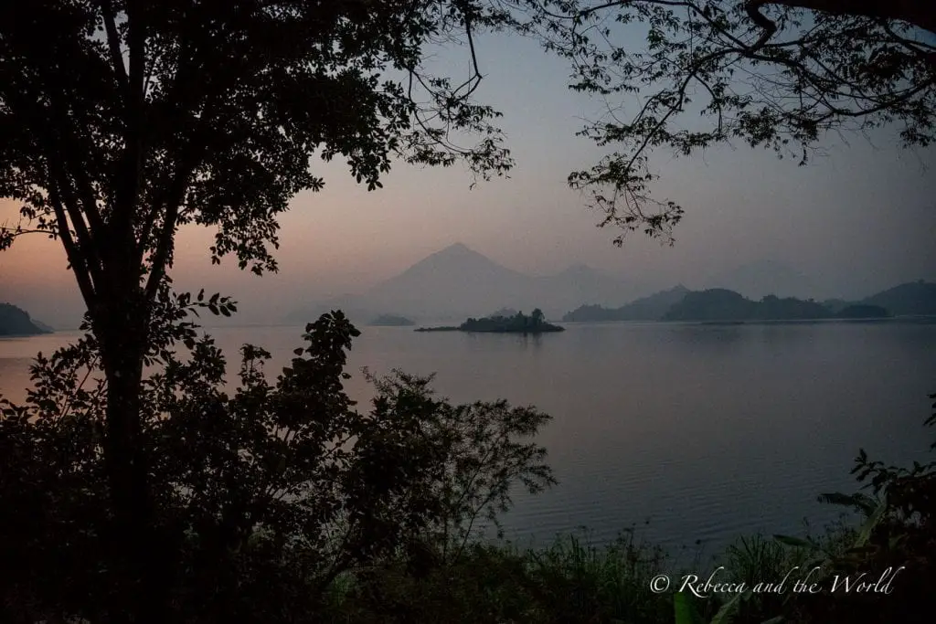 A serene dusk scene beside a lake with trees framing a view of distant hills and a hazy sky reflecting on the calm water. The view of Lake Mutanda from Mutanda Lake Resort in Uganda.